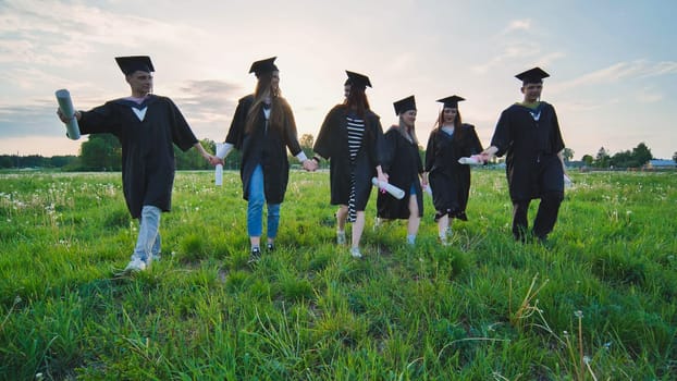 Six graduates in robes walk against the backdrop of the sunset