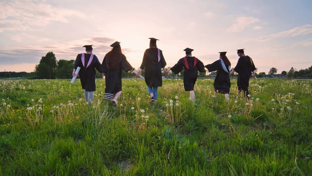 Six graduates in robes walk against the backdrop of the sunset