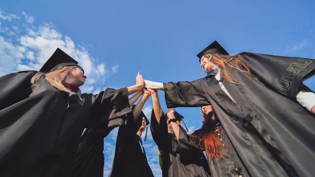 Team of college or university students celebrating graduation. Group of happy successful graduates in academic hats and robes standing in circle and putting their hands together