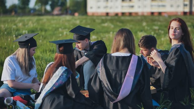 Graduates in black suits eating pizza in a city meadow