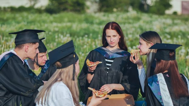 Graduates in black suits eating pizza in a city meadow