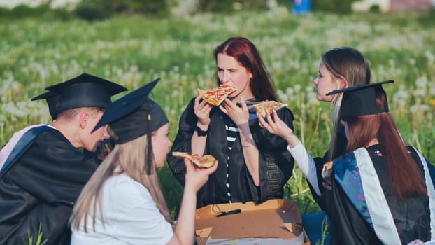 Graduates in black suits eating pizza in a city meadow