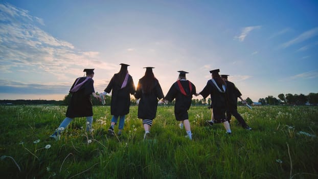 Six graduates in robes walk against the backdrop of the sunset