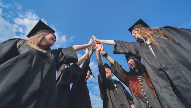 Team of college or university students celebrating graduation. Group of happy successful graduates in academic hats and robes standing in circle and putting their hands together.