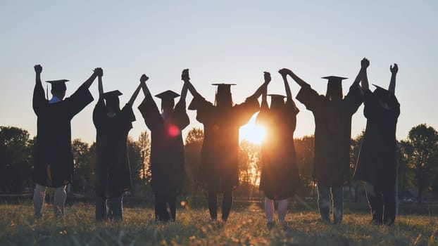 College graduates in robes holding hands at sunset