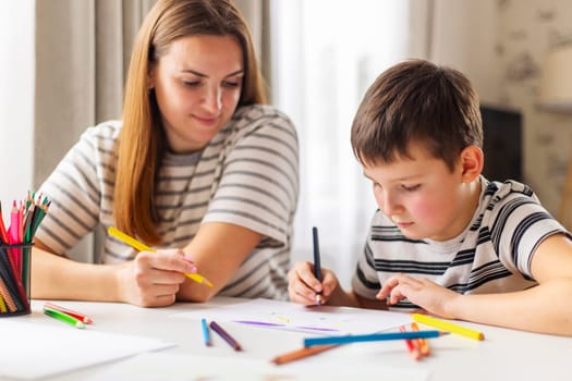 A focused young boy draws with colorful pencils while his smiling mother watches, sharing a creative moment.