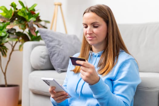 Focused woman in a blue hoodie using a smartphone and credit card to shop online from the comfort of her living room.