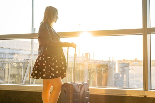 A young woman, dressed in a black jacket and polka-dot dress, stands with a suitcase, waiting to board at the airport during sunset.