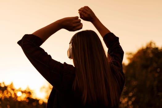 Back view of a woman enjoying music with headphones during a serene sunset, capturing a moment of relaxation.