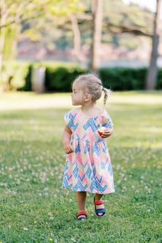 Little girl with an apple in her hand walks on a green lawn, looking away. High quality photo