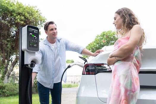 Happy and lovely couple with eco-friendly conscious recharging electric vehicle from EV charging station. EV car technology utilized as alternative transportation for future sustainability. Synchronos