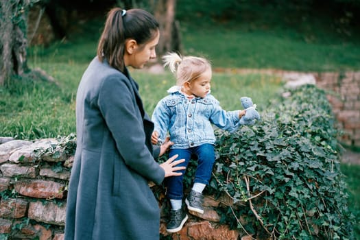 Little girl sits on a stone fence in the park, playing with a soft toy next to a standing mother. High quality photo