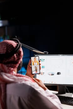 Over-the-shoulder shot of Islamic man conversing with people on his digital mobile device. Detailed view of a male Muslim person sitting at the table using his smartphone for a video conference call.