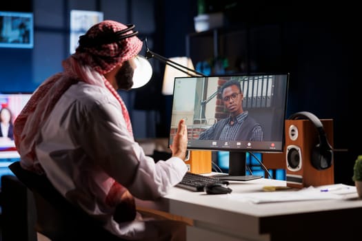 Male African American and Middle Eastern business owners participated in internet video conferencing. Muslim guy dressed in traditional attire listens intently to his coworker on his desktop computer.