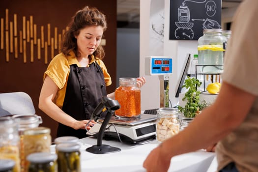 Woman at checkout counter in zero waste shop selling nutritious lentils in jar to customer with green living lifestyle. Client buys freshly harvested food in local neighborhood store