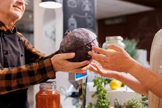 Older man at checkout counter in zero waste shop selling farm grown cabbage to vegan customers with green living lifestyle. Hipster couple buying groceries in local neighborhood store, close up