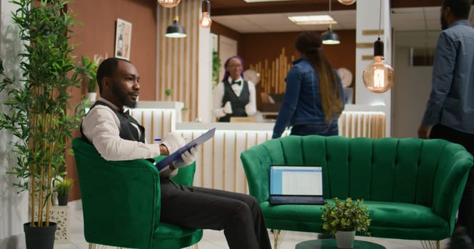 In lobby, employee in tuxedo reviews inventory of bookings. Hotel administrator working as lodging manager and dealing with room reservations on papers. Bellboy handles list of guests.