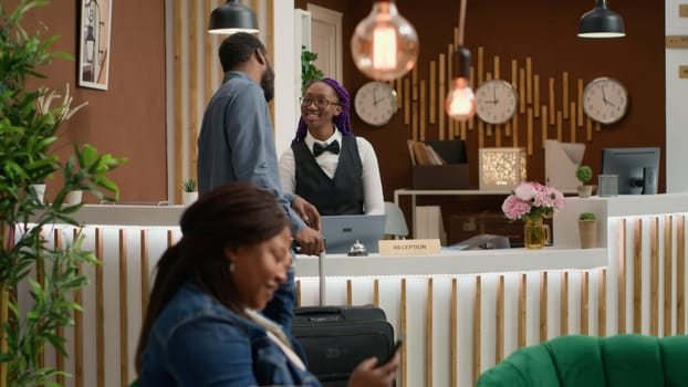 Traveller asks for service and room card at reception, ringing bell to call for receptionist at front desk. African american man with luggage requesting access for hotel accommodation. Handheld shot.