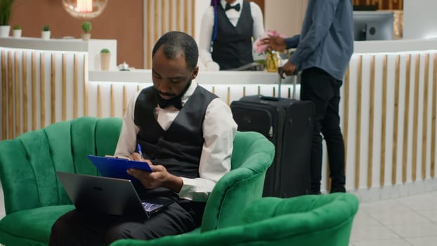 Tourism industry employee. Bellboy entering data document forms on papers in lounge area, hotel concierge making entry in daily luggage movement register, using clipboard notes.