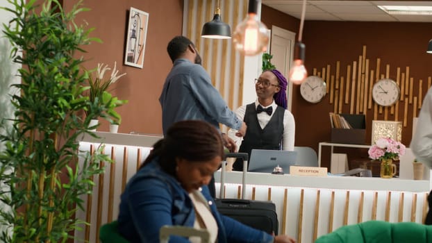 Hotel guest getting access room card after check in process at reception, meeting with front desk staff asking about ammenities. African american man travelling on holiday at resort. Handheld shot.