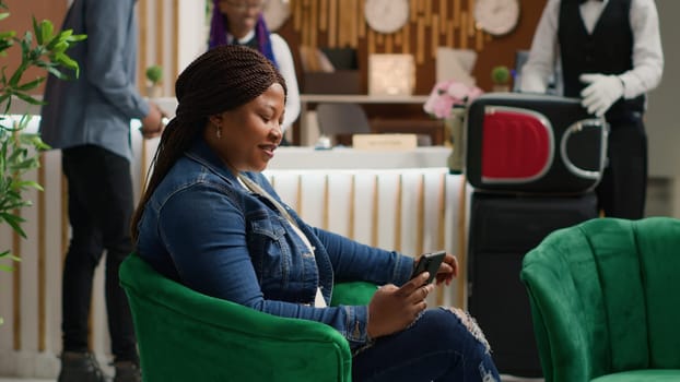 Tourist browsing online page on phone as she sits in lounge area at hotel, using smartphone app to read social media texts. African american woman travelling on vacation. Handheld shot.