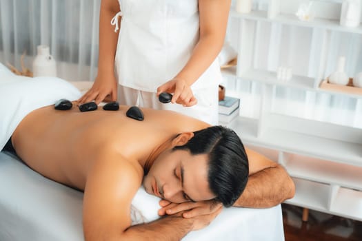 Hot stone massage at spa salon in luxury resort with day light serenity ambient, blissful man customer enjoying spa basalt stone massage glide over body with soothing warmth. Quiescent