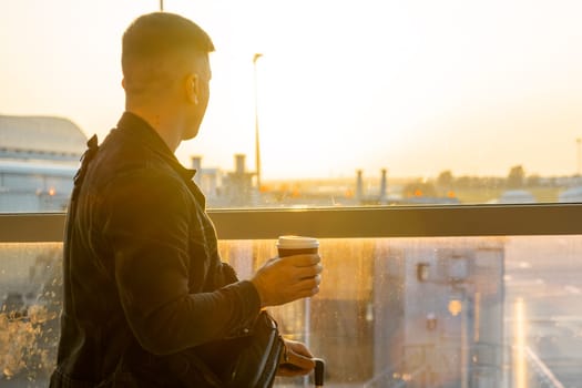 A young man, holding a cup of coffee is ready for boarding with his suitcase.