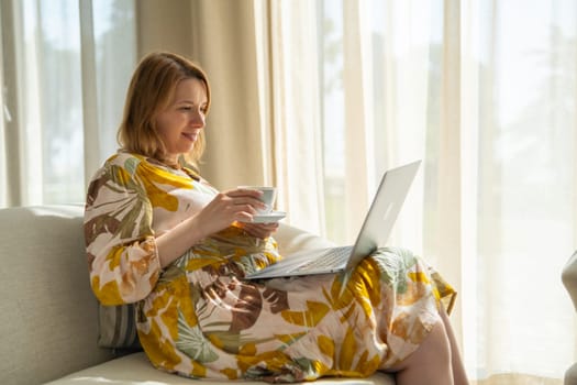 Pregnant woman, in a flowered dress, drinking tea and using a laptop sitting in the hotel hall.