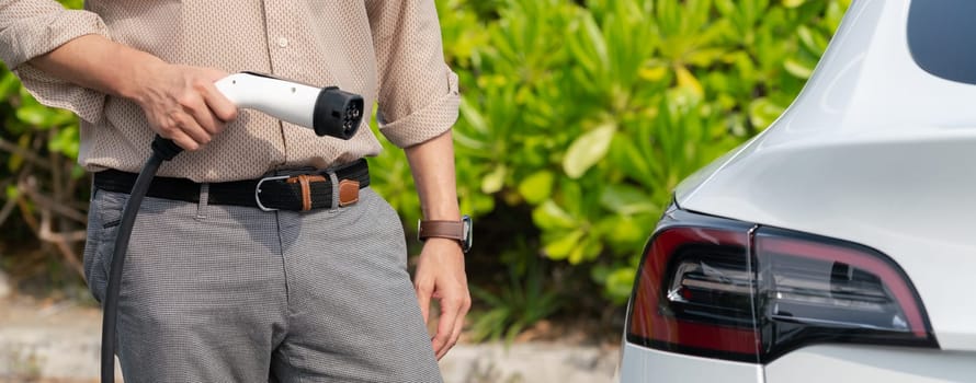 Young man recharge electric car's battery from charging station in outdoor green city park. Rechargeable EV car for sustainable environmental friendly urban travel. Panorama Expedient