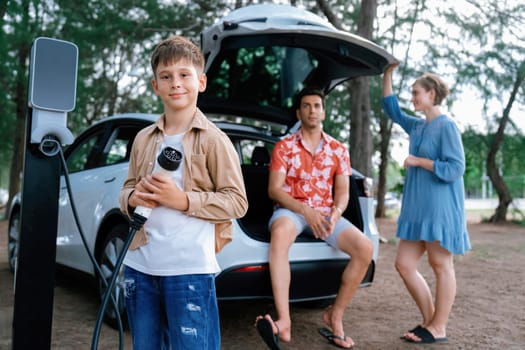 Little boy holding EV charger and point at camera with his family sitting on the trunk in background. Road trip travel with alternative energy charging station for eco-friendly car concept. Perpetual