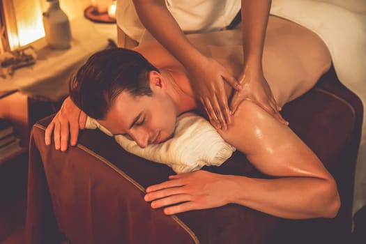 Caucasian man customer enjoying relaxing anti-stress spa massage and pampering with beauty skin recreation leisure in warm candle lighting ambient salon spa at luxury resort or hotel. Quiescent