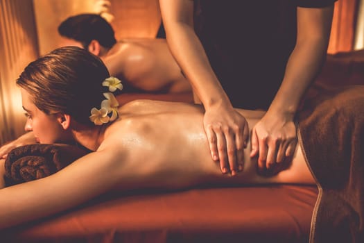 Caucasian couple customer enjoying relaxing anti-stress spa massage and pampering with beauty skin recreation leisure in warm candle lighting ambient salon spa at luxury resort or hotel. Quiescent