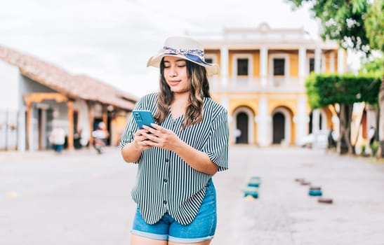 Beautiful young tourist woman in hat using phone on the street. Latin tourist girl texting with phone on the street
