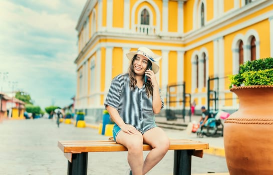 The girl is smiling wearing a hat while sitting on a bench calling on the phone with the background out of focus