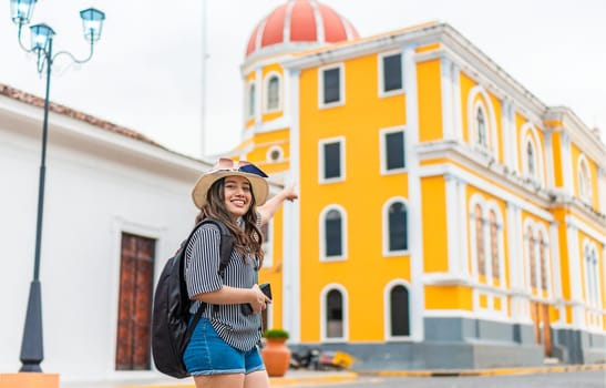 Smiling traveler woman with backpack pointing to a cathedral in a tourist square. Happy tourist girl exploring the city of Granada, Nicaragua
