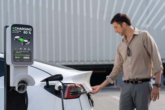 Young man put EV charger to recharge electric car's battery from charging station in city commercial parking lot. Rechargeable EV car for sustainable environmental friendly urban travel. Expedient