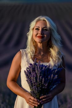 Blonde woman poses in lavender field at sunset. Happy woman in white dress holds lavender bouquet. Aromatherapy concept, lavender oil, photo session in lavender.