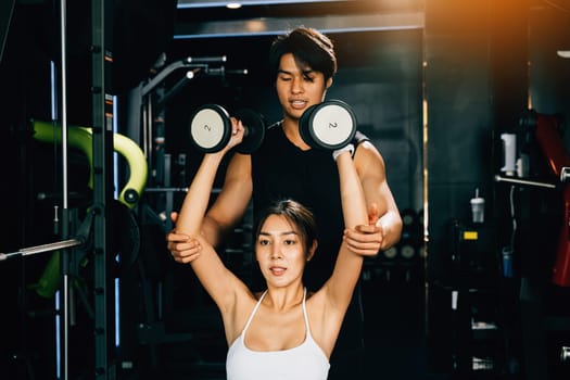 Fit Asian woman lifting dumbbells with a smile, supported by her personal trainer in a gym filled with modern exercise equipment, technique of exercise in gym dark gym background
