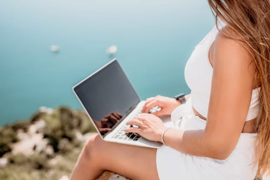 Successful business woman in yellow hat working on laptop by the sea. Pretty lady typing on computer at summer day outdoors. Freelance, travel and holidays concept.