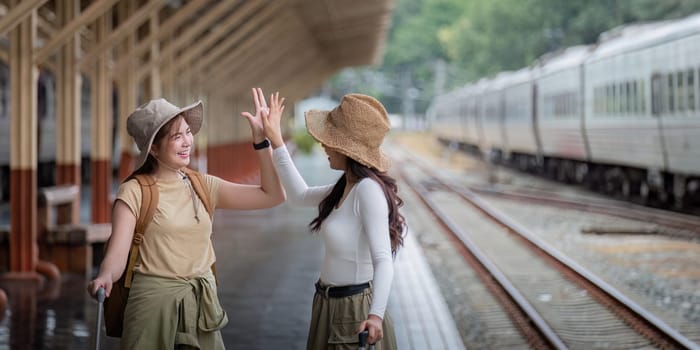 Two women are happy while traveling at the train station. tour concept. Two asian women best friend travel high five on train timetable in railway station.