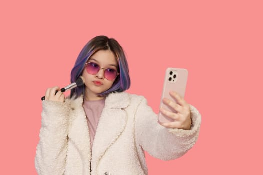 Stylish Teenager Girl, In Modern Pink Glasses, Uses Mobile Phone As Mirror While Applying Makeup With Brush Against Pink Background. Fashionable And Trendy Teenager Embracing Contemporary Beauty Practices.High quality photo