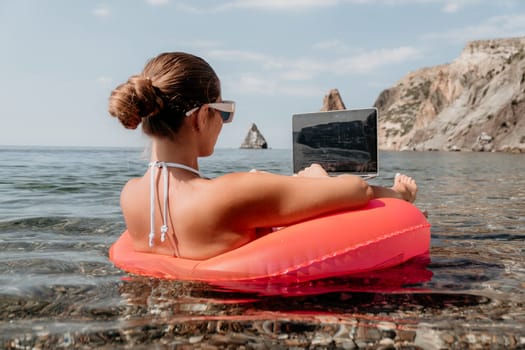 Woman freelancer works on laptop swimming in sea on pink inflatable ring. Pretty lady typing on computer while floating in the sea on inflatable donut at sunset. Freelance, remote work on vacation