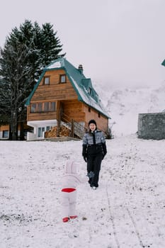 Small child walks towards his laughing mother along a snow-covered hill near a wooden cottage. Back view. High quality photo