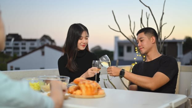 Smiling woman and man talking and clinking glasses at rooftop party during sunset.