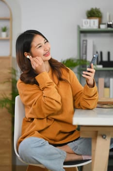 Cheerful young woman listen to music in wireless earphones and using mobile phone in living room.
