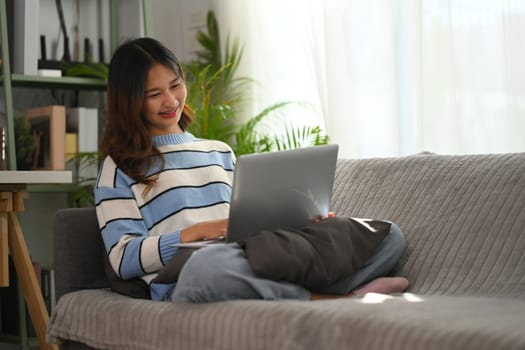 Portrait of smiling young woman sitting on grey sofa and using laptop.