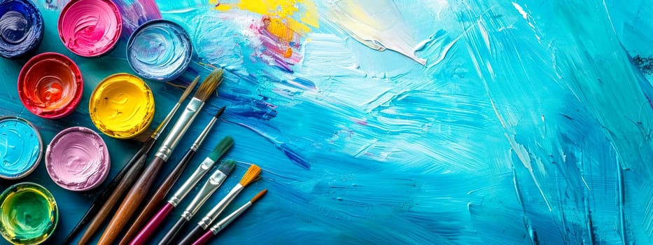 A colorful array of open paint pots and various-sized brushes on a vivid turquoise painted canvas background, showcasing creativity and the art-making process. banner with copy space