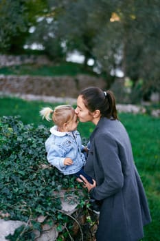 Smiling mom kisses the forehead of a little girl sitting on a stone fence in the garden. High quality photo
