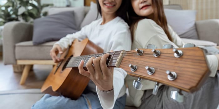 Lesbian couple lean against each other and sit and play guitar relaxed and happy together at home..
