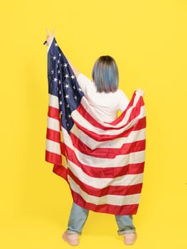 Victory Sign By Girl Covered With American Flag, Patriotic Pride, Cultural Diversity, And Freedom. Teenager Isolated On Yellow Backdrop With Copy Space For Your Text. Full-Height Backside View . High quality photo